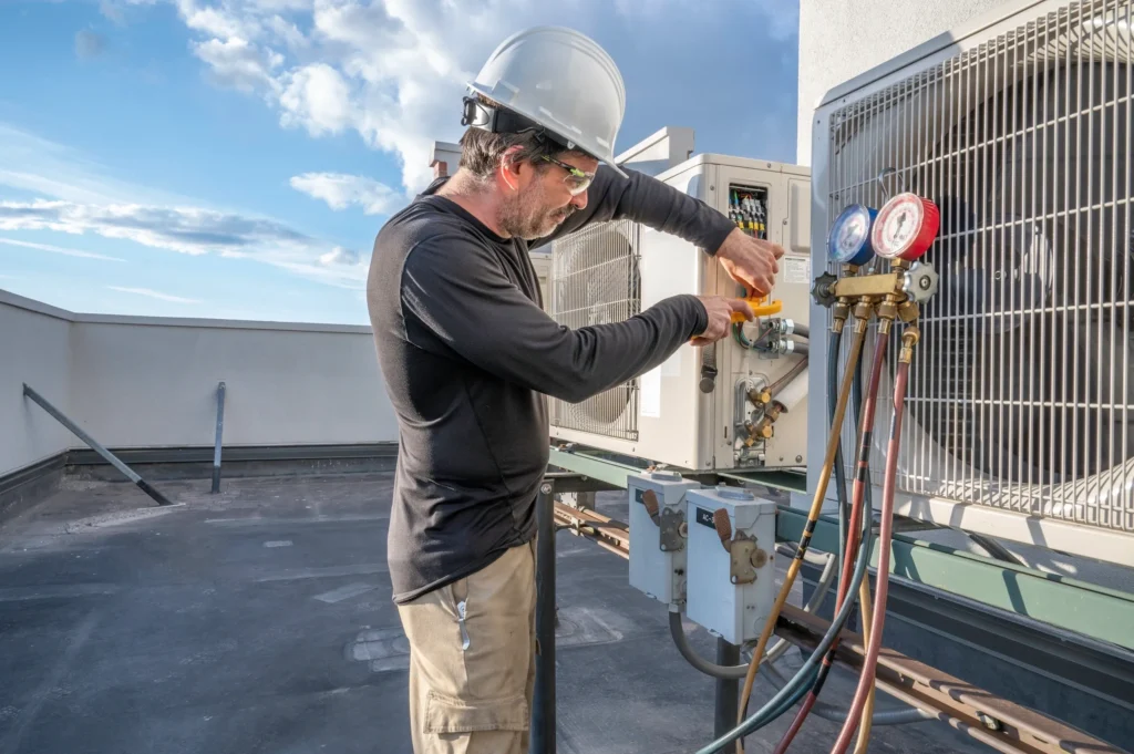A technician in a hard hat connects refrigerant gauges to a rooftop air conditioning unit.