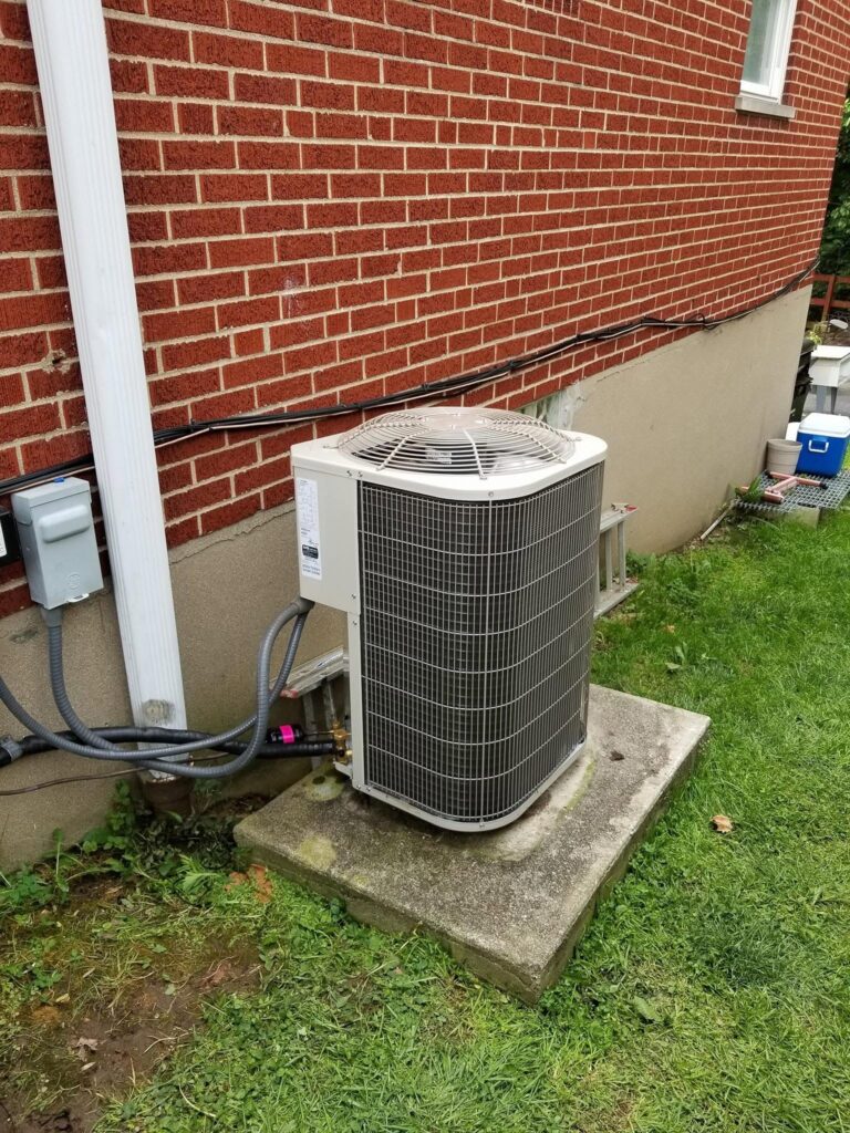 An outdoor central air conditioner condenser on a concrete pad with open space around it beside a brick house.